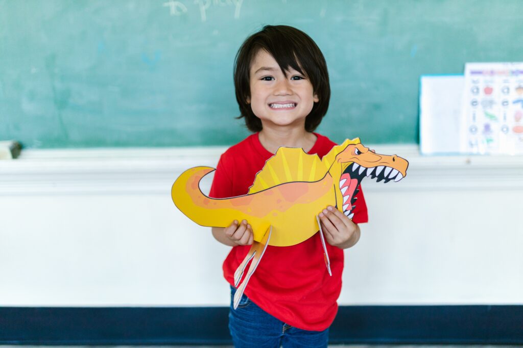 Little boy holding dinosaur toy in classroom in front of chalkboard.