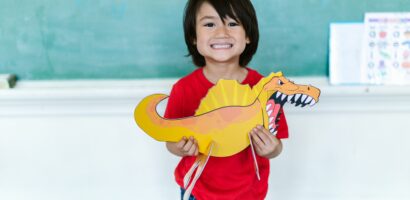 Little boy holding dinosaur toy in classroom in front of chalkboard.